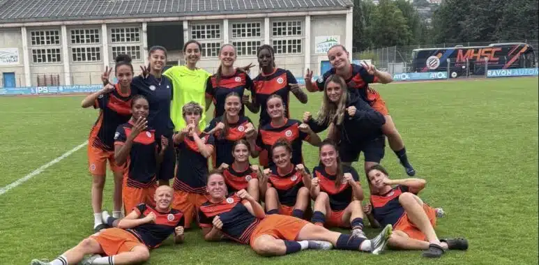 Screenshot | AllezPaillade.com Group of young female soccer players posing on a grass field in navy and orange uniforms, smiling and making victory signs after a match.