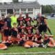 Group of young female soccer players posing on a grass field in navy and orange uniforms, smiling and making victory signs after a match.