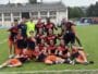 Group of young female soccer players posing on a grass field in navy and orange uniforms, smiling and making victory signs after a match.