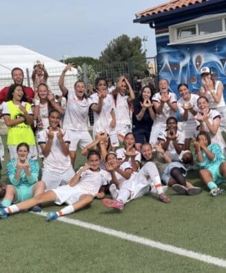 Group of soccer players and coaches posing for a team photo on a green field, many in white uniforms with a few in yellow bibs, cheering and making hand gestures.