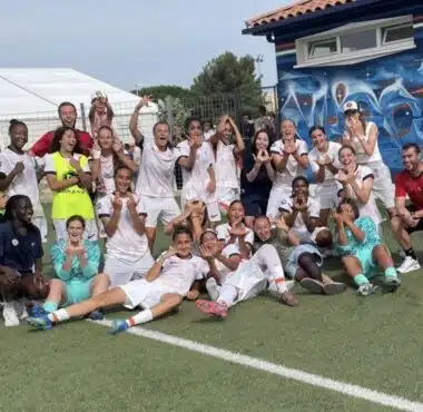 Group of soccer players and coaches posing for a team photo on a green field, many in white uniforms with a few in yellow bibs, cheering and making hand gestures.