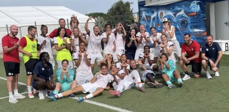 Group of soccer players and coaches posing for a team photo on a green field, many in white uniforms with a few in yellow bibs, cheering and making hand gestures.