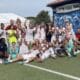 Group of soccer players and coaches posing for a team photo on a green field, many in white uniforms with a few in yellow bibs, cheering and making hand gestures.