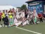 Group of soccer players and coaches posing for a team photo on a green field, many in white uniforms with a few in yellow bibs, cheering and making hand gestures.