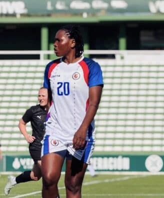 Soccer player in a white jersey with number 20 on a green field, standing with others in the background at a stadium.