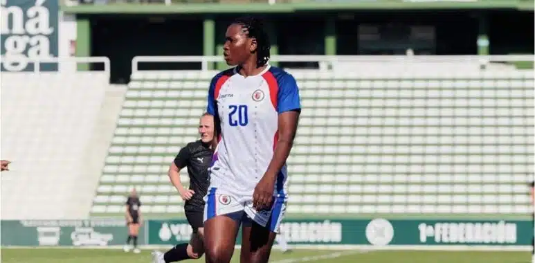 Soccer player in a white jersey with number 20 on a green field, standing with others in the background at a stadium.