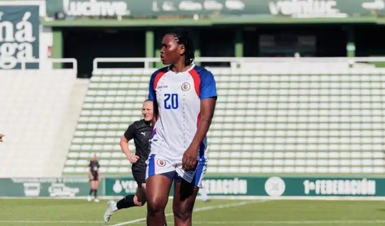 Soccer player in a white jersey with number 20 on a green field, standing with others in the background at a stadium.