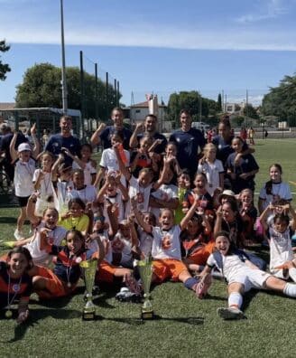Screenshot | AllezPaillade.com Youth soccer team posing with trophies and medals after a match on a sunny field. Players and coaches celebrate together.