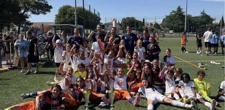 Screenshot | AllezPaillade.com Youth soccer team posing with trophies and medals after a match on a sunny field. Players and coaches celebrate together.
