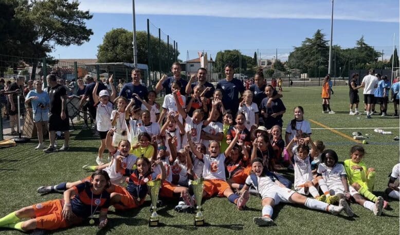 Screenshot | AllezPaillade.com Youth soccer team posing with trophies and medals after a match on a sunny field. Players and coaches celebrate together.
