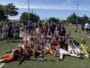 Youth soccer team posing with trophies and medals after a match on a sunny field. Players and coaches celebrate together.