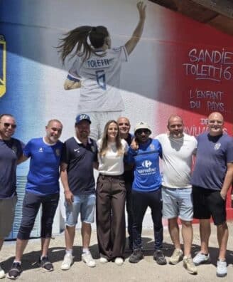 Group of ten people posing together in front of a colorful mural with a football club crest on the wall behind them on a sunny day.