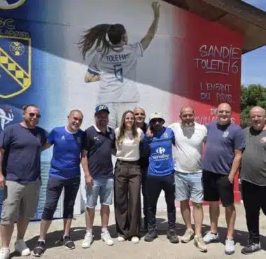 Group of ten people posing together in front of a colorful mural with a football club crest on the wall behind them on a sunny day.