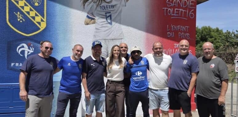Screenshot | AllezPaillade.com Group of ten people posing together in front of a colorful mural with a football club crest on the wall behind them on a sunny day.