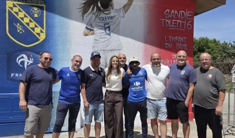 Screenshot | AllezPaillade.com Group of ten people posing together in front of a colorful mural with a football club crest on the wall behind them on a sunny day.
