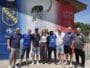 Group of ten people posing together in front of a colorful mural with a football club crest on the wall behind them on a sunny day.
