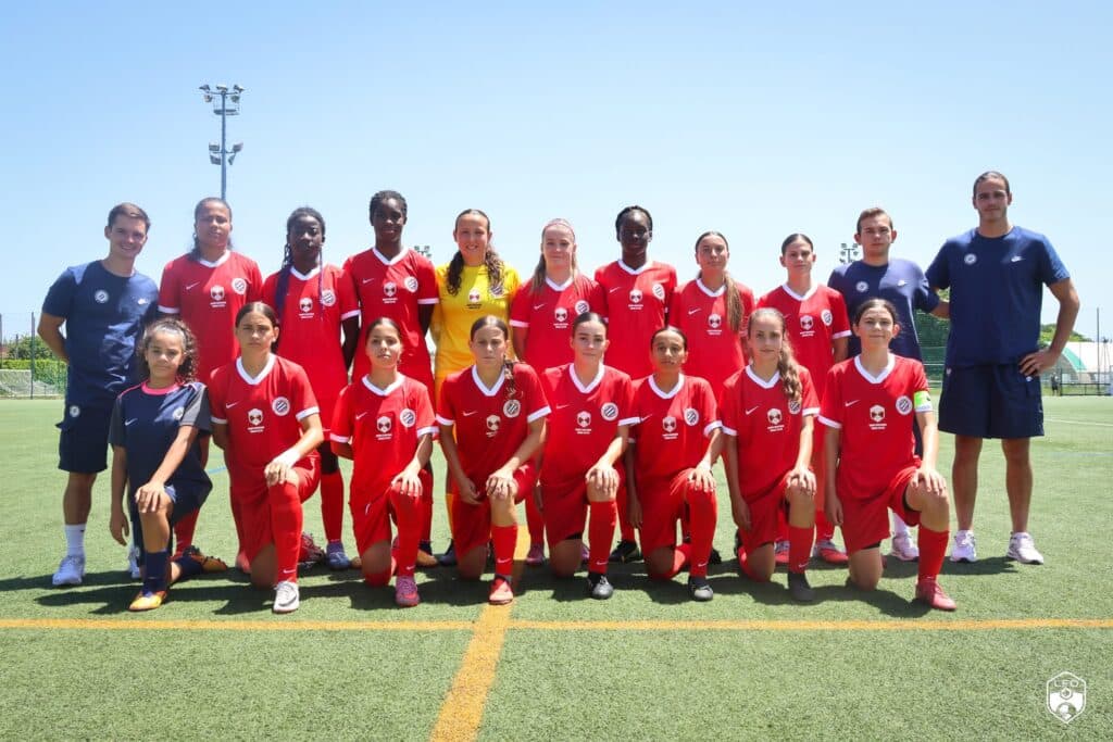 IMG_5049 | AllezPaillade.com Group of youth soccer players in red uniforms posing for a team photo on a sunny field, with two coaches in blue at the ends and a yellow goalie in the center.
