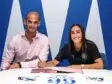 Two professionals signing documents at a white table; man in a pink shirt, woman in black polo, blue backdrop.