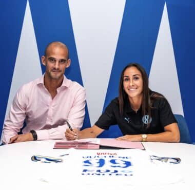 Two professionals signing documents at a white table; man in a pink shirt, woman in black polo, blue backdrop.