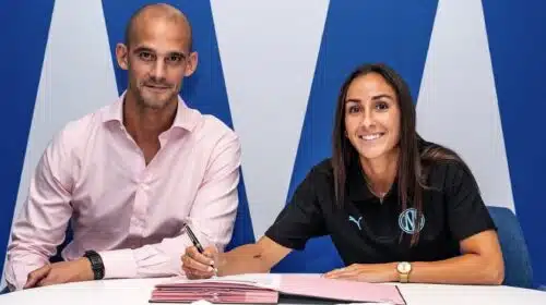 Two professionals signing documents at a white table; man in a pink shirt, woman in black polo, blue backdrop.