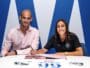 Two professionals signing documents at a white table; man in a pink shirt, woman in black polo, blue backdrop.