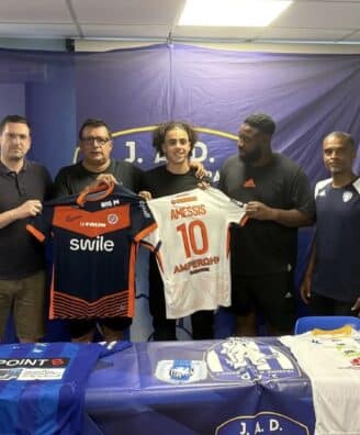 Five men stand side by side indoors, holding two soccer jerseys&mdash;orange/black and white with the name AMESSIS and number 10&mdash;in front of a blue backdrop with a banner at the top.