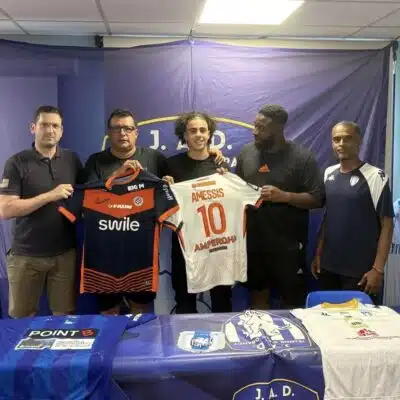 Five men stand side by side indoors, holding two soccer jerseys&mdash;orange/black and white with the name AMESSIS and number 10&mdash;in front of a blue backdrop with a banner at the top.
