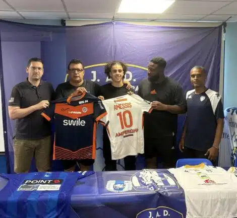Five men stand side by side indoors, holding two soccer jerseys&mdash;orange/black and white with the name AMESSIS and number 10&mdash;in front of a blue backdrop with a banner at the top.
