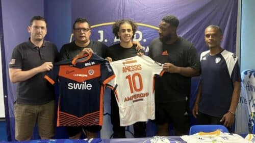 Five men stand side by side indoors, holding two soccer jerseys&mdash;orange/black and white with the name AMESSIS and number 10&mdash;in front of a blue backdrop with a banner at the top.