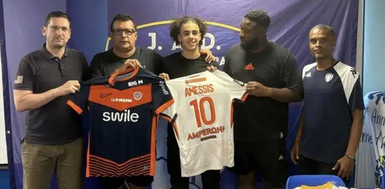 Screenshot | AllezPaillade.com Five men stand side by side indoors, holding two soccer jerseys—orange/black and white with the name AMESSIS and number 10—in front of a blue backdrop with a banner at the top.