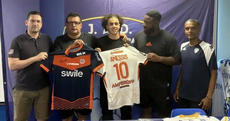 Screenshot | AllezPaillade.com Five men stand side by side indoors, holding two soccer jerseys—orange/black and white with the name AMESSIS and number 10—in front of a blue backdrop with a banner at the top.