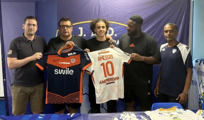 Five men stand side by side indoors, holding two soccer jerseys&mdash;orange/black and white with the name AMESSIS and number 10&mdash;in front of a blue backdrop with a banner at the top.