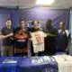 Five men stand side by side indoors, holding two soccer jerseys&mdash;orange/black and white with the name AMESSIS and number 10&mdash;in front of a blue backdrop with a banner at the top.