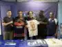 Five men stand side by side indoors, holding two soccer jerseys&mdash;orange/black and white with the name AMESSIS and number 10&mdash;in front of a blue backdrop with a banner at the top.