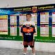 Young boy holding an orange and navy soccer jersey in front of a wall of league schedules and charts.