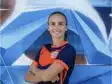 Smiling woman in an orange and navy soccer jersey with arms crossed in front of a blue geometric mural.