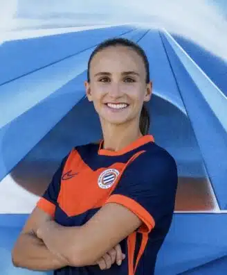 Smiling woman in an orange and navy soccer jersey with arms crossed in front of a blue geometric mural.