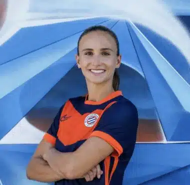 Smiling woman in an orange and navy soccer jersey with arms crossed in front of a blue geometric mural.
