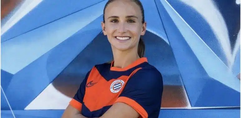 Screenshot | AllezPaillade.com Smiling woman in an orange and navy soccer jersey with arms crossed in front of a blue geometric mural.