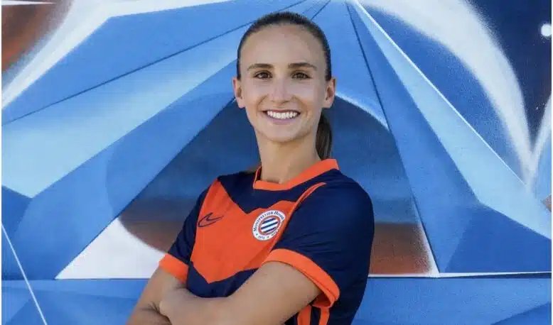 Smiling woman in an orange and navy soccer jersey with arms crossed in front of a blue geometric mural.