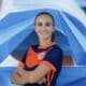 Smiling woman in an orange and navy soccer jersey with arms crossed in front of a blue geometric mural.