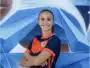 Smiling woman in an orange and navy soccer jersey with arms crossed in front of a blue geometric mural.
