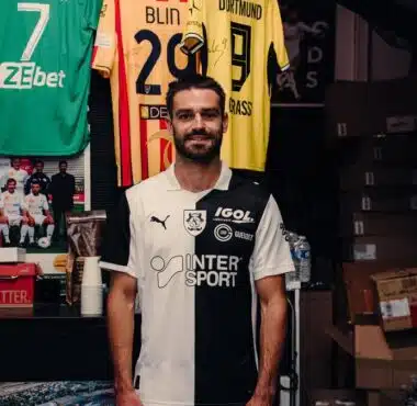 Man in a black-and-white football jersey standing in a team shop with jerseys hanging behind him.