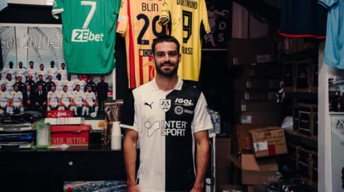 Man in a black-and-white football jersey standing in a team shop with jerseys hanging behind him.