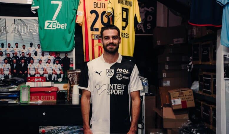 Man in a black-and-white football jersey standing in a team shop with jerseys hanging behind him.