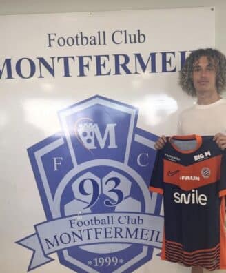 Screenshot | AllezPaillade.com Young man with curly hair posing beside a large Football Club Montfermeil sign, holding a navy and orange Montfermeil football jersey.