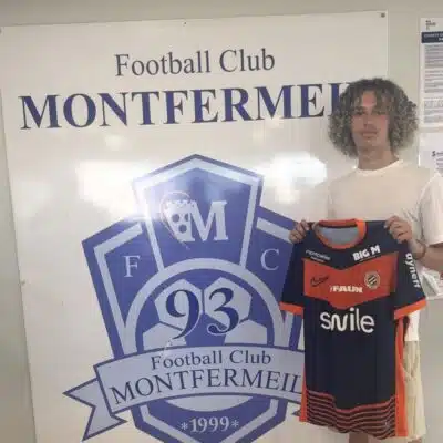 Young man with curly hair posing beside a large Football Club Montfermeil sign, holding a navy and orange Montfermeil football jersey.