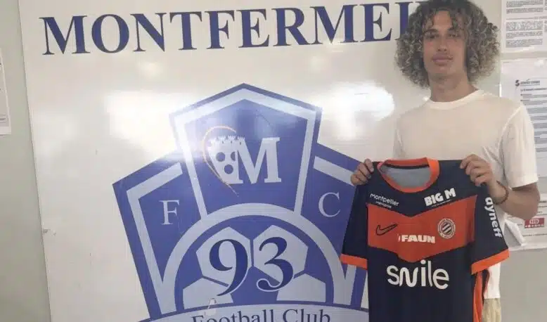 Screenshot | AllezPaillade.com Young man with curly hair posing beside a large Football Club Montfermeil sign, holding a navy and orange Montfermeil football jersey.