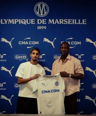 Two men pose with a white CMA CGM football jersey against a blue Olympique de Marseille backdrop with sponsor logos nearby, indoors.
