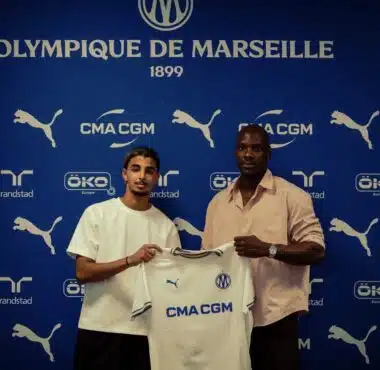 Two men pose with a white CMA CGM football jersey against a blue Olympique de Marseille backdrop with sponsor logos nearby, indoors.